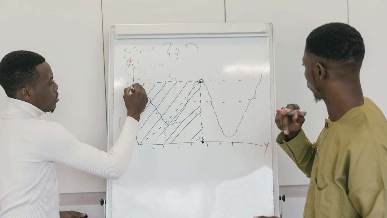 Two men at a whiteboard, one drawing a chart while the other points at it.